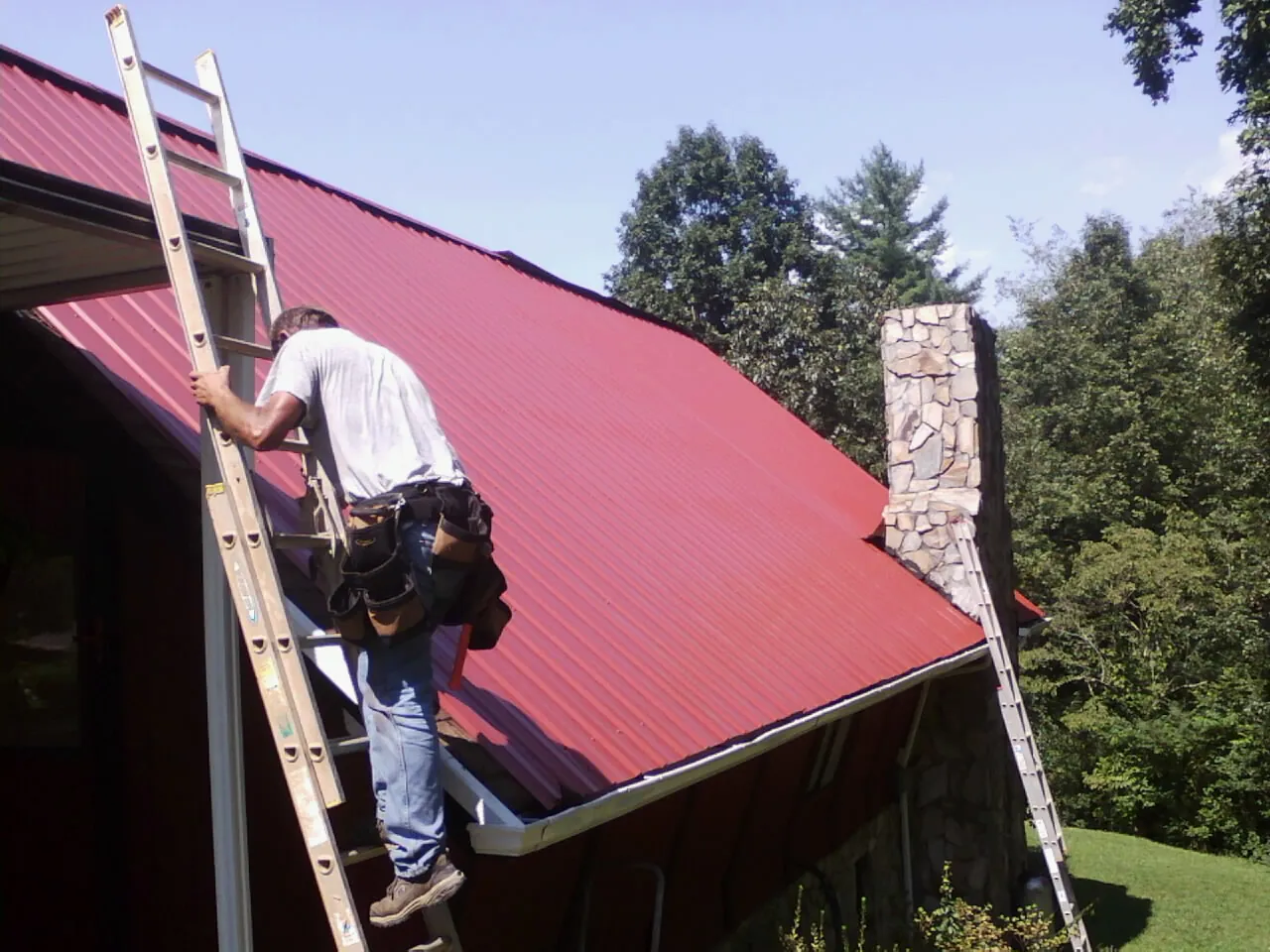 Roofer on a ladder beside a red metal roof and stone chimney — Soffit & Fascia Repair by Roofing Brothers in Sheridan